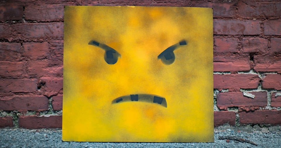 Emoji style painting on a square canvas with angry eyes and a frown against a brick wall.