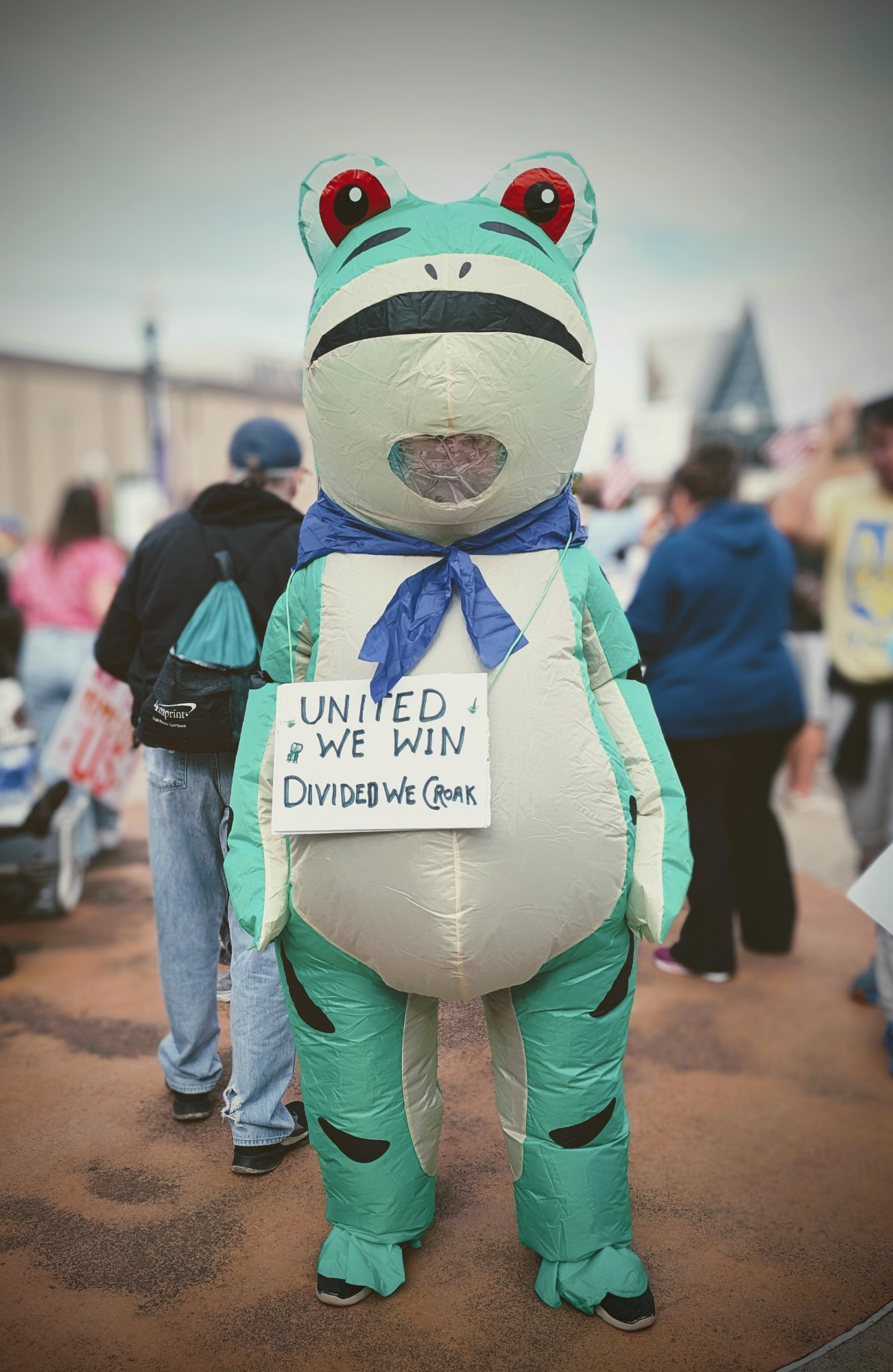 A protester in a frog costume at No Kings 2.0 with a sign around its neck that says United We Win divided we croak.