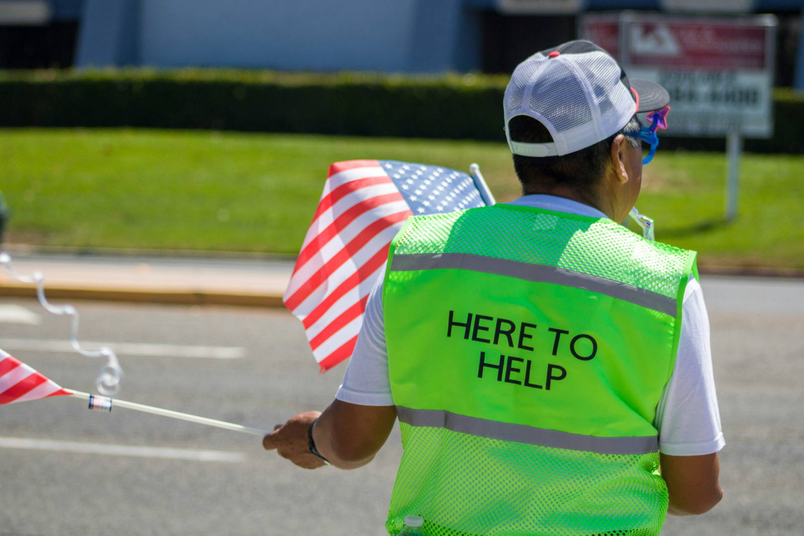 Man in a white cap and a yellow high visibility vest that says Here to Help waving American flags